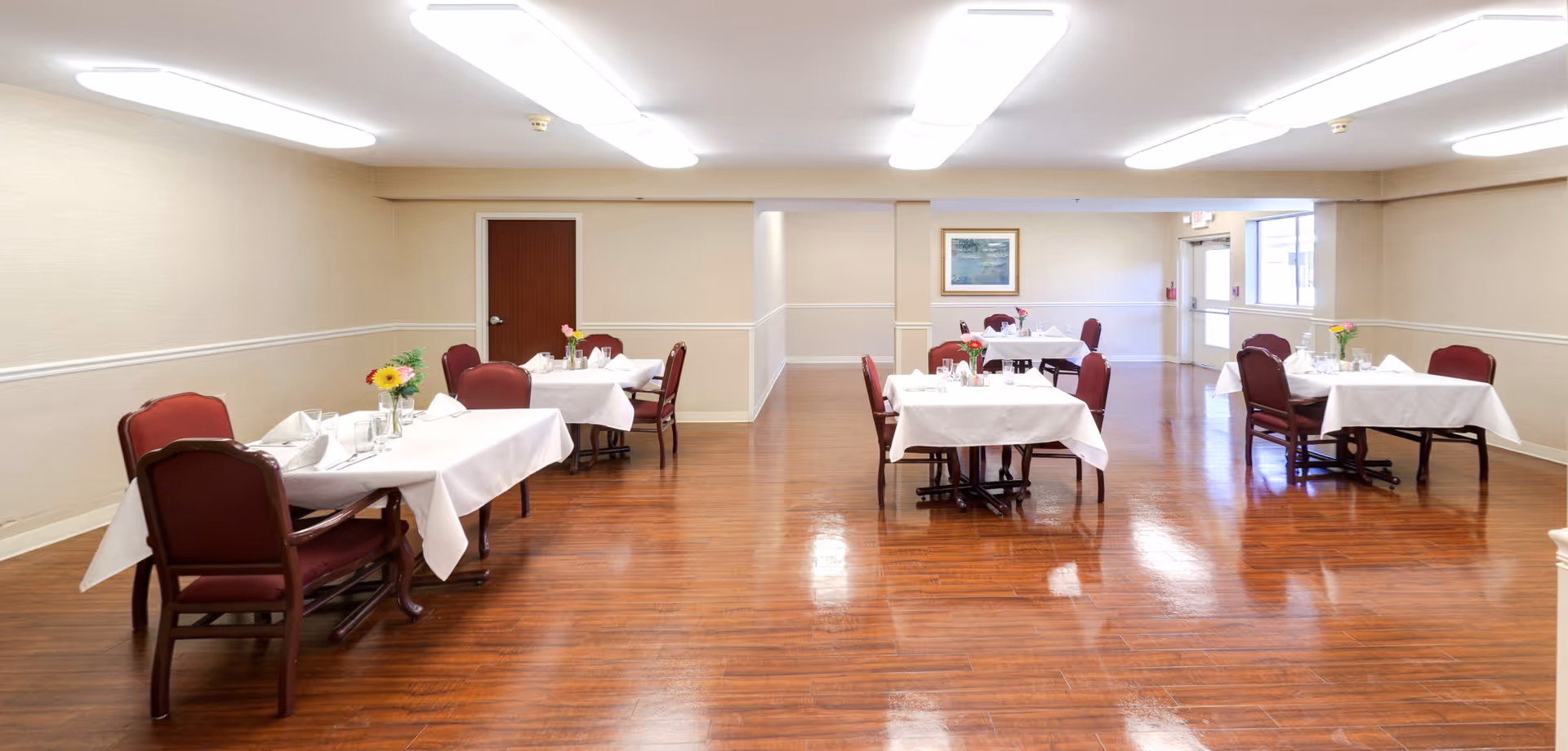 A dining room with several tables covered in white tablecloths, each set with glassware and floral centerpieces. The room has wooden flooring, beige walls, and maroon cushioned chairs. There is a door and a framed picture on the far wall, and a large window letting in natural light.