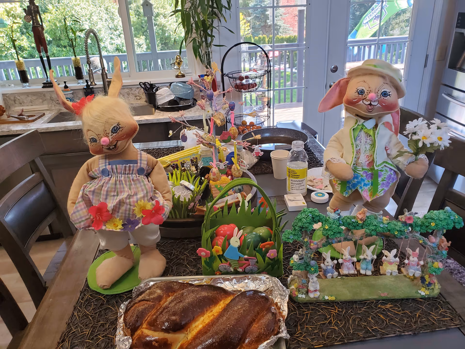 Dining table set with Easter decorations including two large bunny dolls, baskets of colorful eggs and a braided loaf of bread in a home kitchen/dining area.