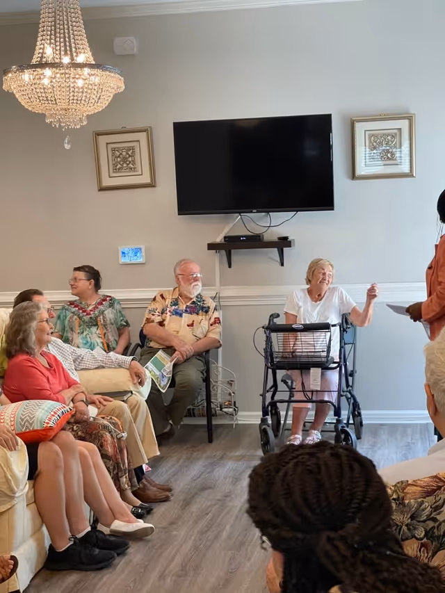 A group of elderly people seated in a living room setting, some on a couch and others on chairs, engaging in a conversation. One elderly woman is using a walker and appears to be speaking to a standing person holding papers. The room has a chandelier, a wall-mounted TV, framed artwork, and wood flooring.