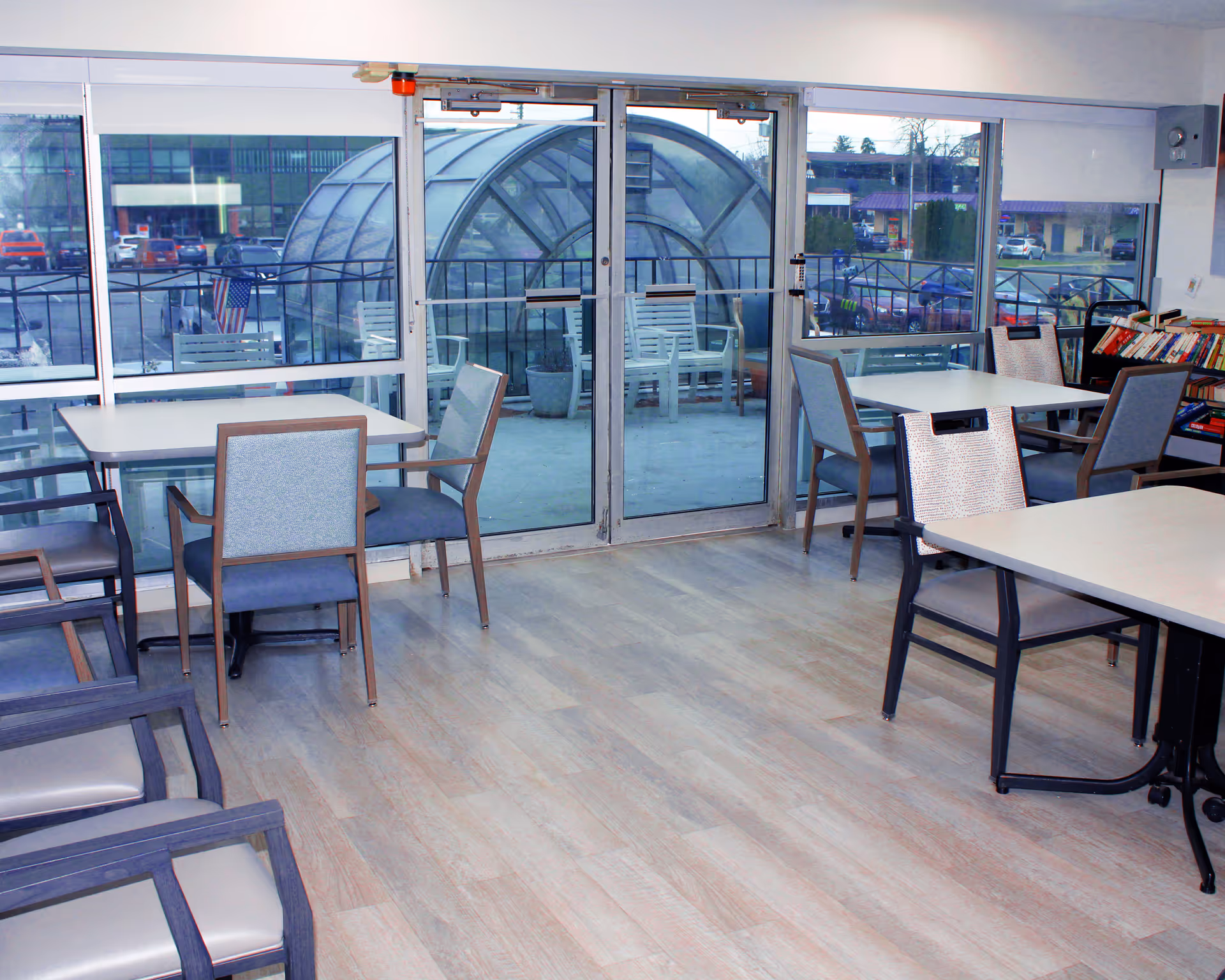 Communal dining/activity room with tables and chairs facing large glass sliding doors overlooking an outdoor seating area and parking lot.