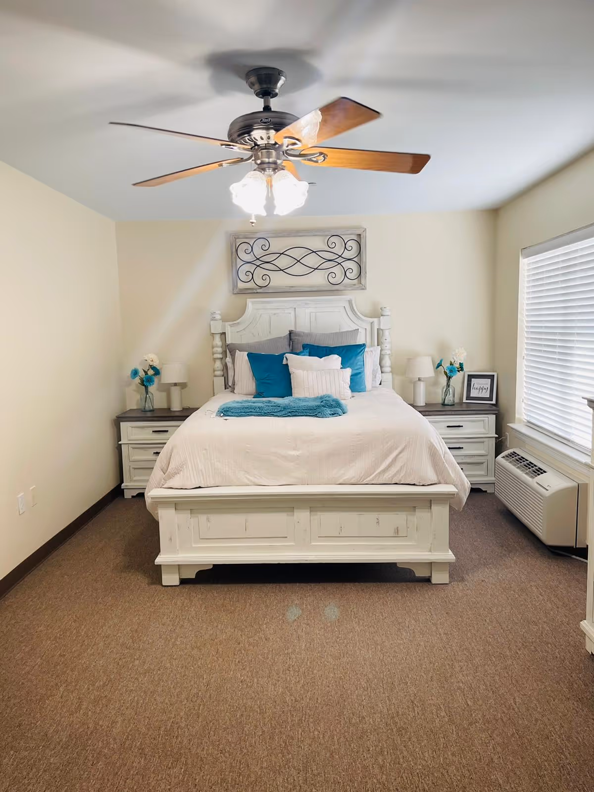 A neatly made bed with white bedding and blue accent pillows in a bedroom. The bed has a white wooden headboard and footboard. There are two matching white nightstands on either side of the bed, each with a lamp and a vase with blue and white flowers. A framed picture is on the right nightstand. A ceiling fan with lights is mounted on the ceiling. A window with white blinds is on the right wall, and a wall-mounted air conditioning unit is below the window. The carpet is brown, and the walls are painted light beige.