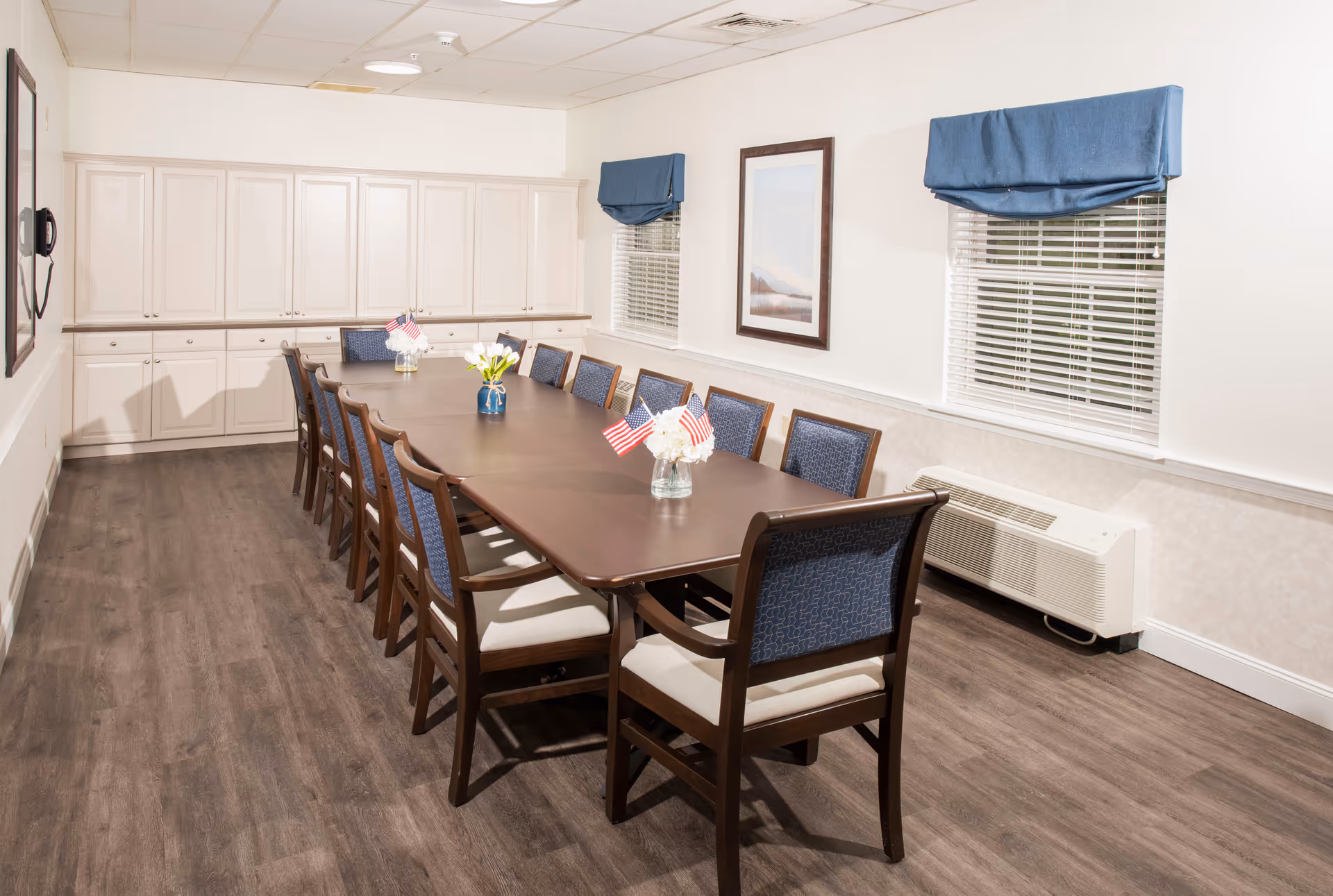 A long rectangular dining table with twelve chairs arranged around it in a well-lit room. The table has small vases with flowers and miniature American flags. The room features wood flooring, white walls with two windows covered by blue valances, a framed picture on the wall, and white cabinetry along the back wall.