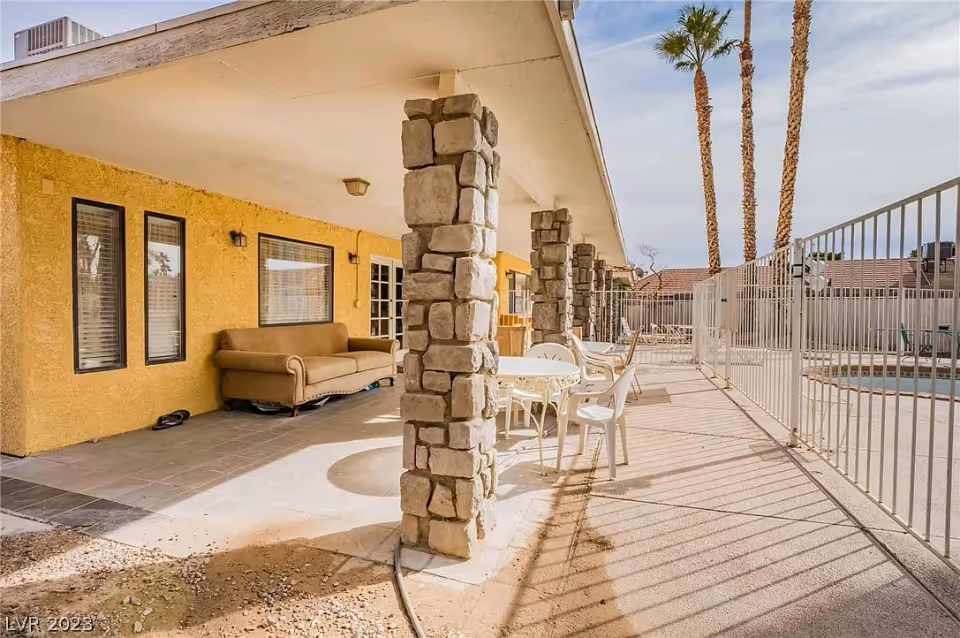 Outdoor covered patio area with stone pillars, a brown couch, white metal table with chairs, and a fenced swimming pool. Palm trees and neighboring houses are visible in the background under a partly cloudy sky.