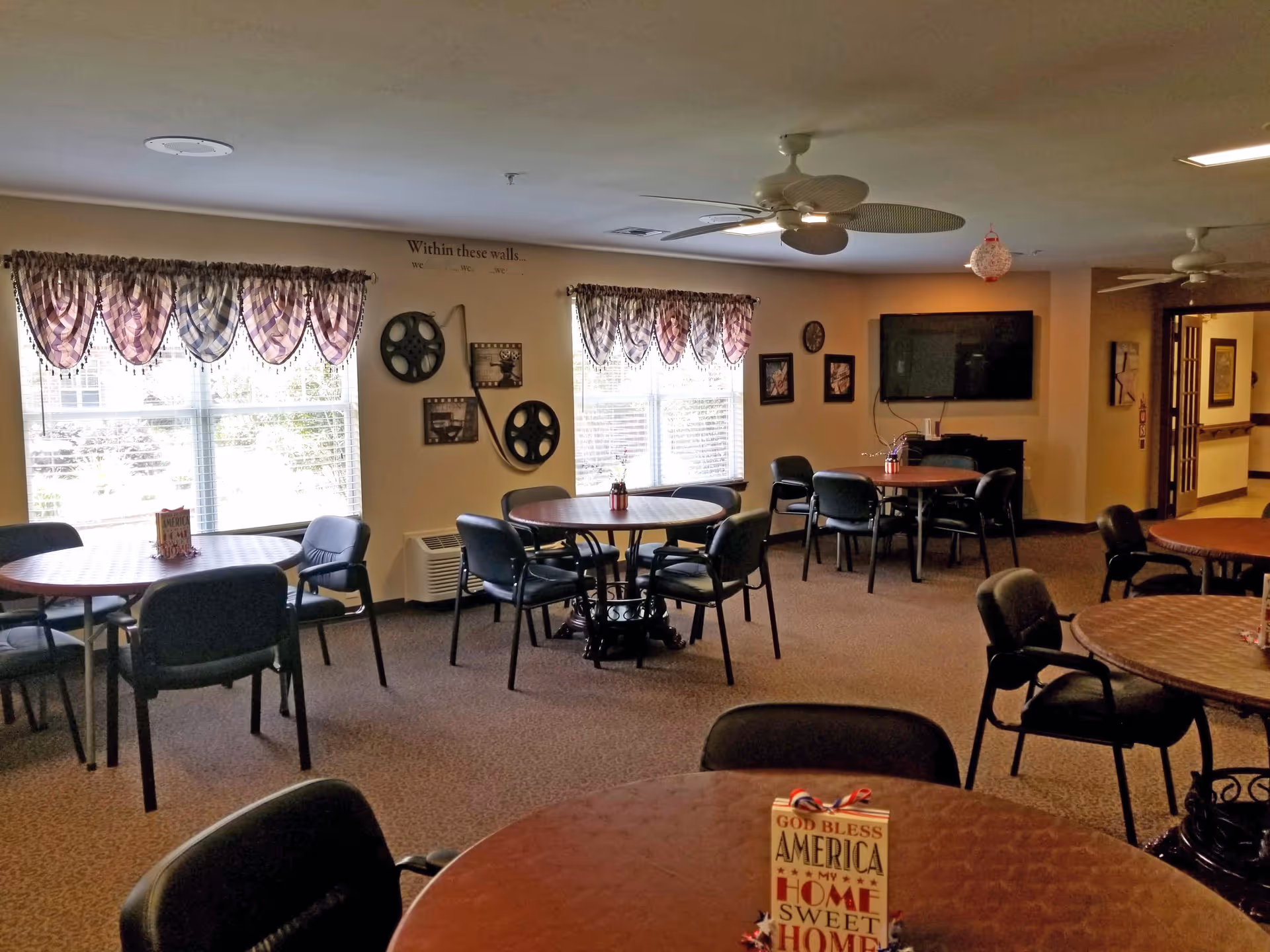 A communal dining/activity room with round tables, chairs, ceiling fans, a wall-mounted TV, and windows with valances.