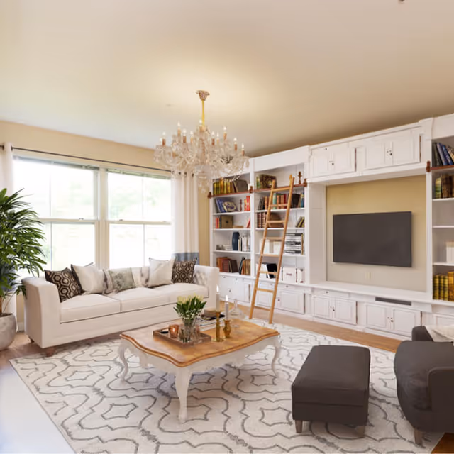 Bright living room with a white sofa, wooden coffee table on a patterned rug, built-in bookshelves with a rolling ladder, and a wall-mounted TV.