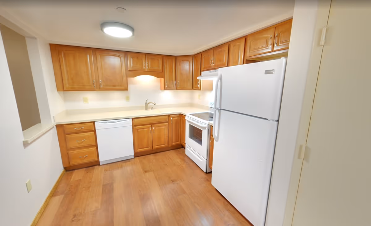 A clean kitchen with wooden cabinets, a white refrigerator, a white stove with an oven, a white dishwasher, and a sink under a cabinet with under-cabinet lighting. The floor is wooden and the walls are light-colored.