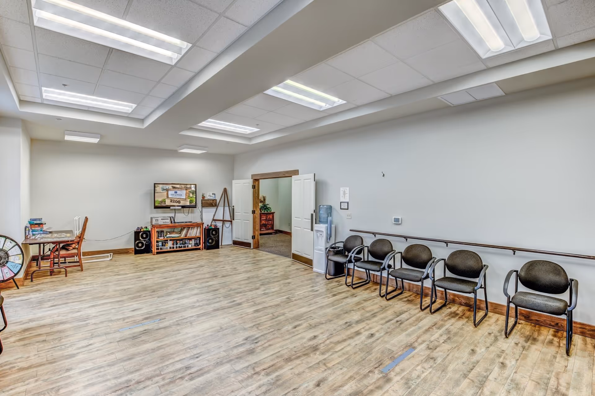 Bright communal activity room with a row of chairs along one wall, a TV and bookshelf, a game table, and a water cooler.