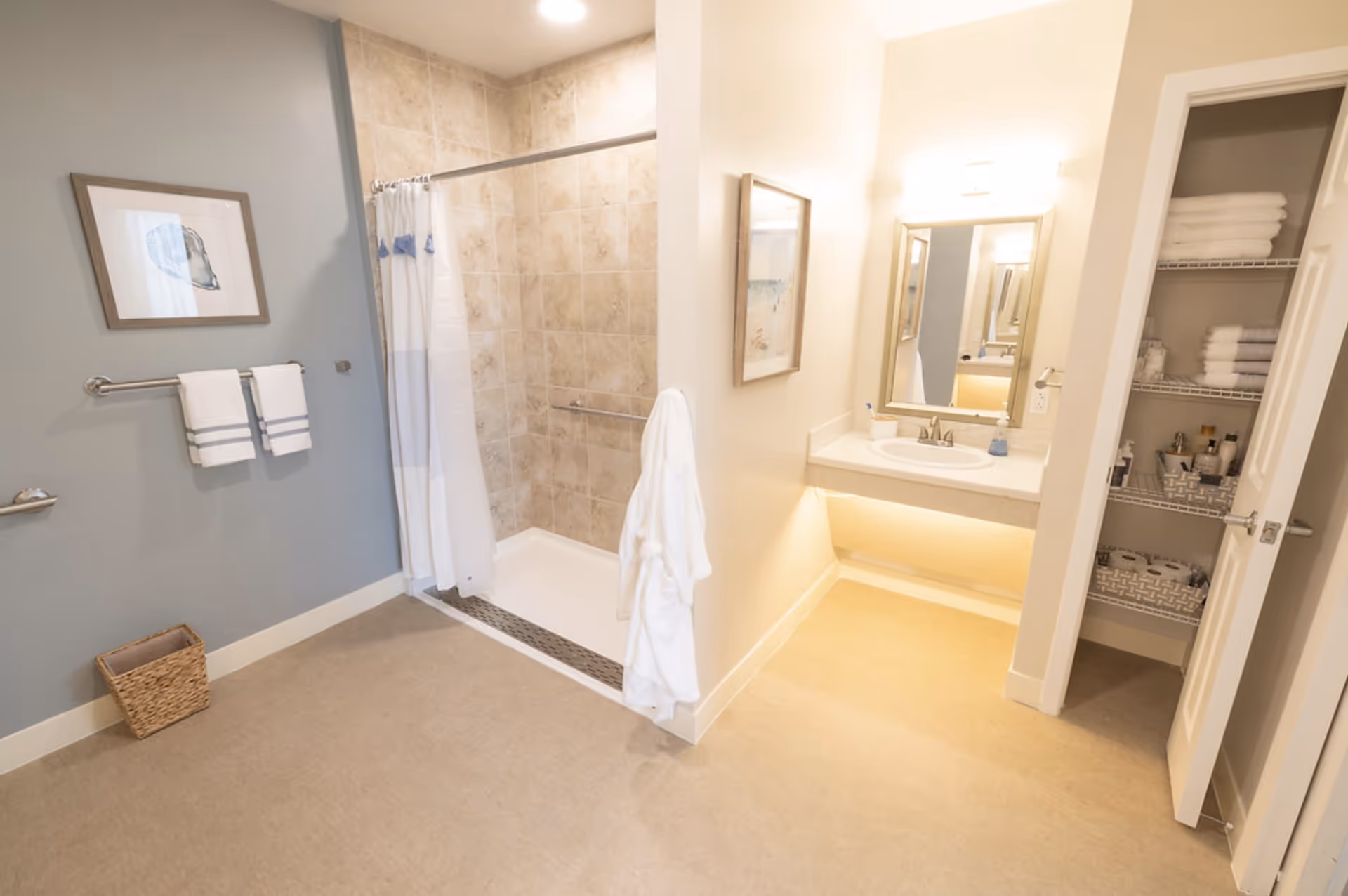 A clean and spacious bathroom featuring a walk-in shower with beige tiled walls and a white shower curtain. There is a white robe hanging on the corner wall, a vanity with a sink and mirror, and an open linen closet stocked with towels and toiletries. The walls are painted in light colors, and there is a framed picture on the wall.