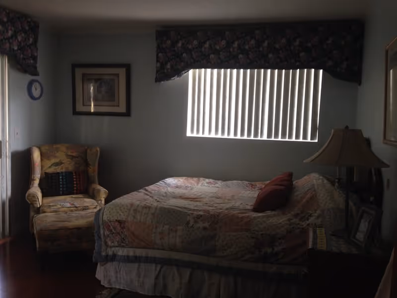 Dimly lit bedroom with a bed, patterned armchair, bedside lamp, and a window with vertical blinds.