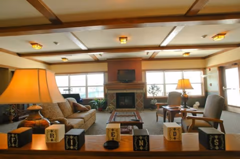 A spacious and warmly lit living room with a beige sofa, two armchairs, a coffee table, and a fireplace with a TV mounted above it. The room has large windows letting in natural light, wooden ceiling beams, and two table lamps providing additional lighting. Decorative blocks spelling 'FRIENDS' are placed on a wooden ledge in the foreground.