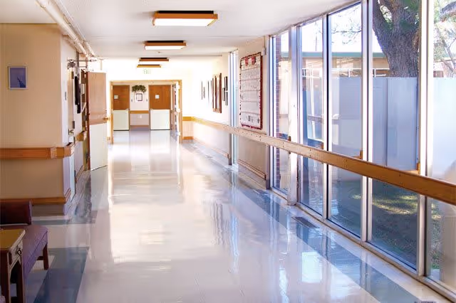 A bright hallway in a healthcare facility with large windows on the right side letting in natural light. The hallway has a shiny tiled floor, handrails along the walls, and doors at the far end. There is a bulletin board on the right wall and some framed pictures on the left wall.