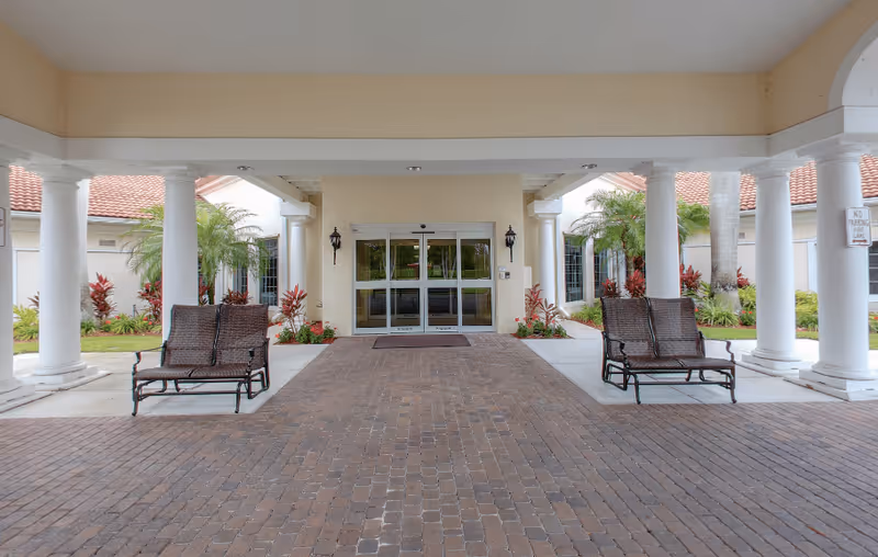 Entrance area of a senior living facility with a covered driveway supported by white columns. Two brown wicker benches are placed on either side of the driveway. The entrance features glass double doors with wall-mounted lantern-style lights on each side. There are plants and small palm trees along the sides near the building walls.