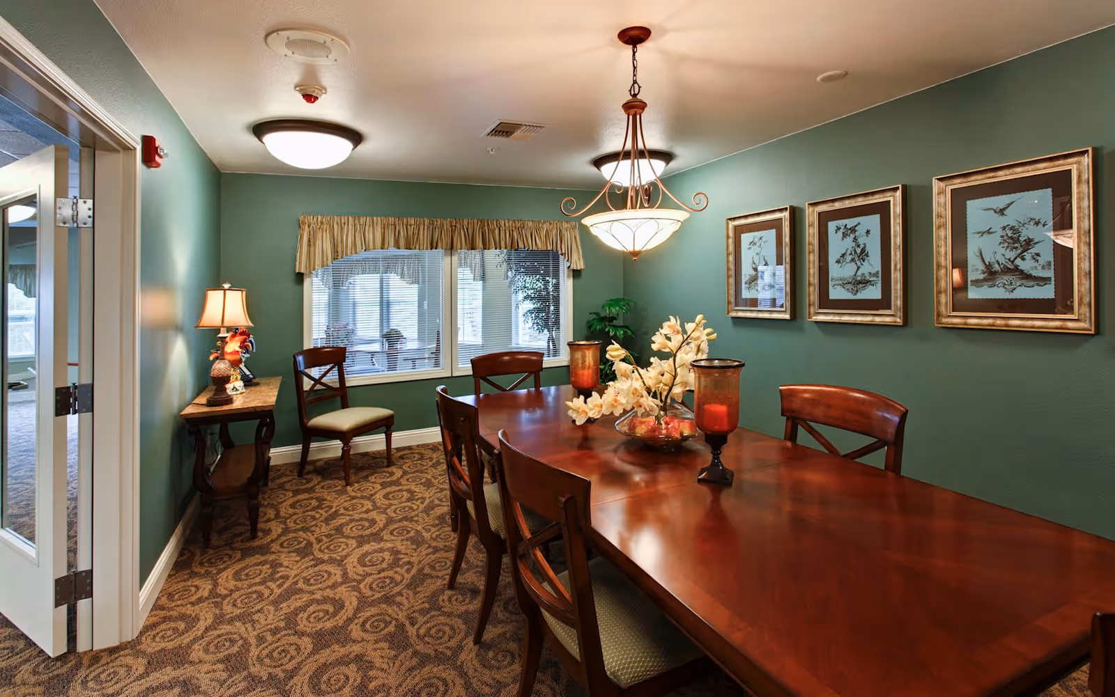 A dining room with a long wooden table surrounded by chairs. The table is decorated with a floral centerpiece and two red candle holders. The walls are painted green and adorned with three framed pictures of birds. There is a window with blinds and a valance, a small side table with a lamp, and a chandelier hanging from the ceiling.