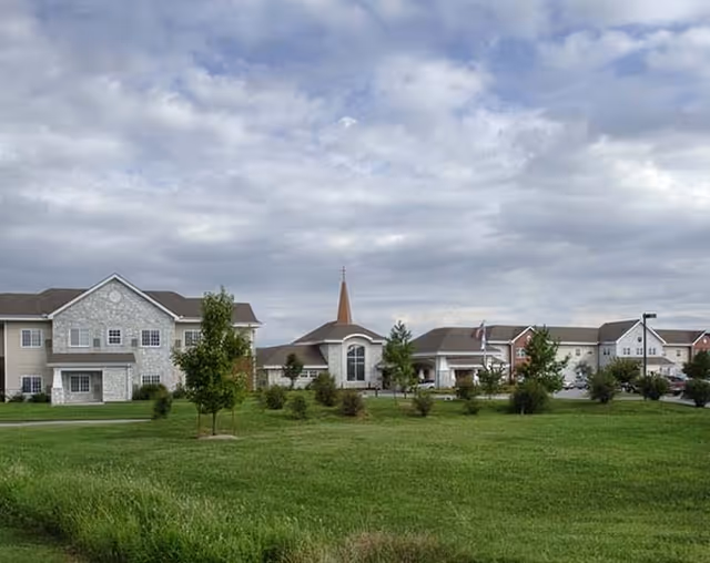 Exterior view of a senior living complex with connected buildings, a central chapel-like section with a steeple, and lawns and trees under a cloudy sky.