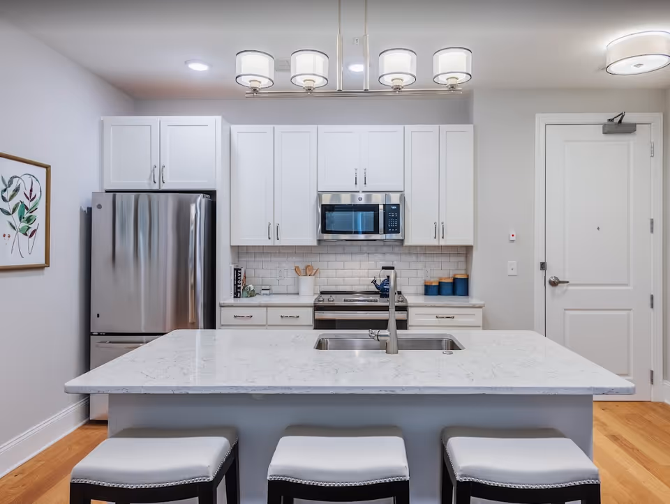 Bright modern kitchen featuring a marble island with a sink and three stools, white cabinets, and stainless steel appliances.