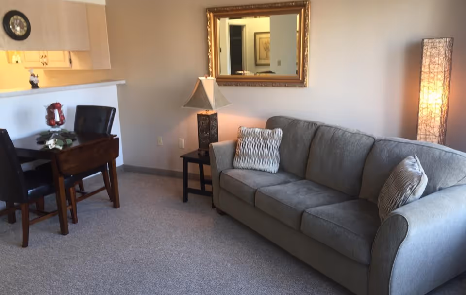 A cozy living room area featuring a gray upholstered sofa with two patterned pillows, a side table with a decorative lamp, a tall floor lamp with a woven design, a wall-mounted mirror with a gold frame, and a small dining table with two dark chairs near a kitchen counter.