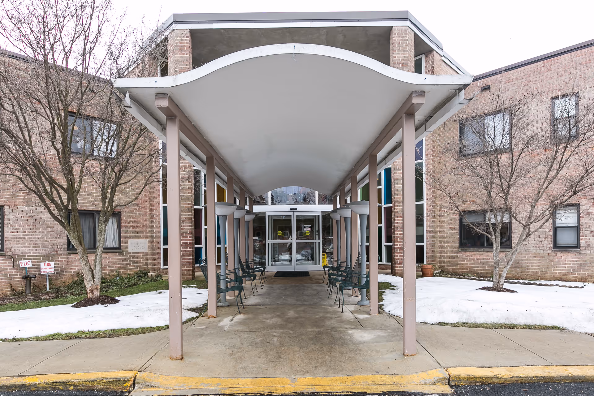 Entrance to a brick building with a covered walkway supported by columns. There are benches along the walkway and leafless trees on either side with patches of snow on the ground. The building has large windows and double glass doors at the entrance.