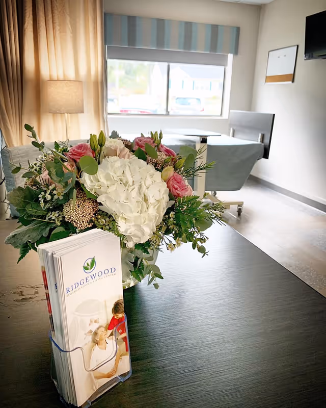 A bright room with a large window showing a view outside. In the foreground, there is a dark wooden table with a bouquet of flowers featuring white hydrangeas and pink roses, alongside a brochure holder containing Ridgewood Rehabilitation & Living Center pamphlets. In the background, there is a hospital-style bed and a floor lamp next to beige curtains.