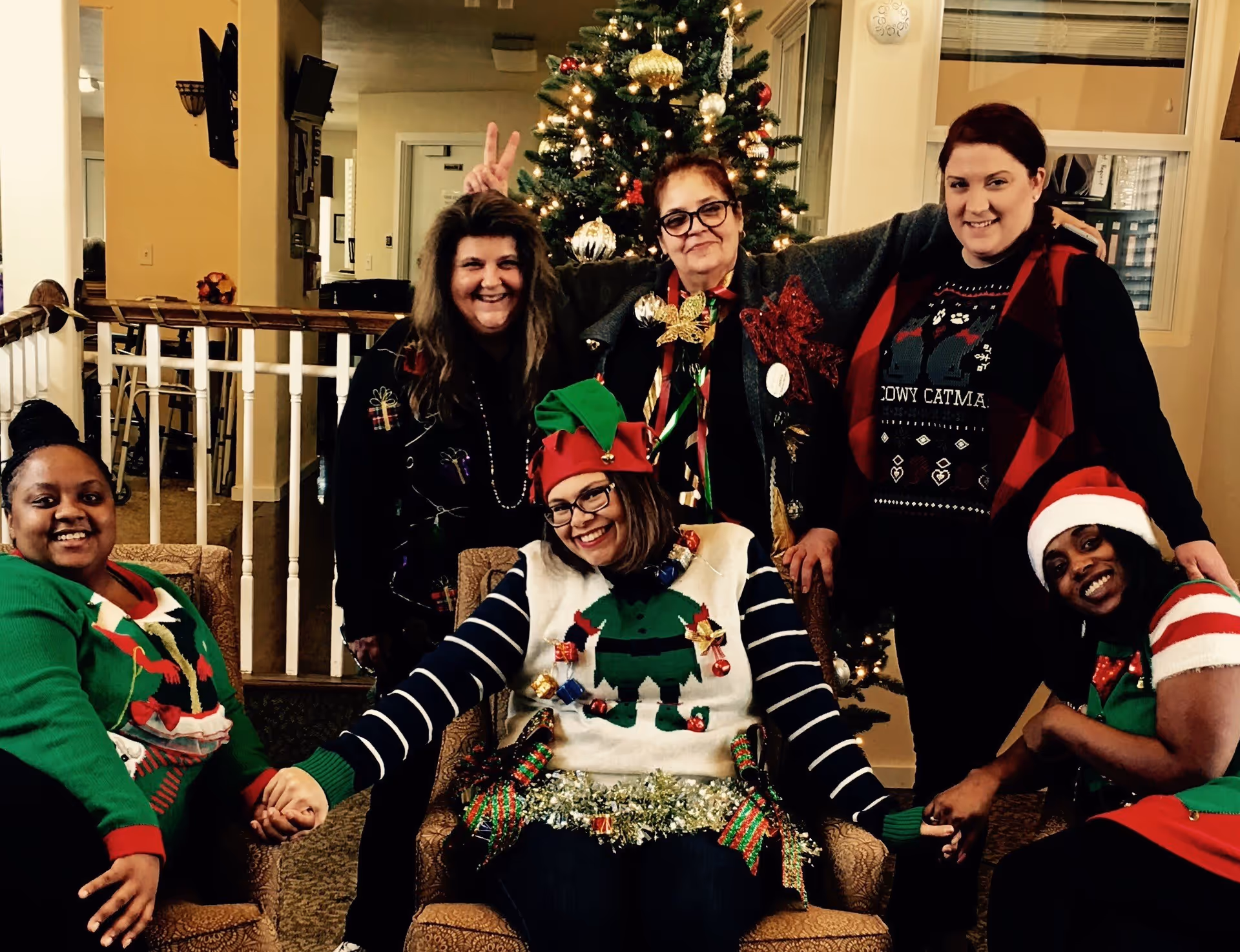 Six people wearing festive holiday sweaters and hats pose and smile in a seating area in front of a decorated Christmas tree.