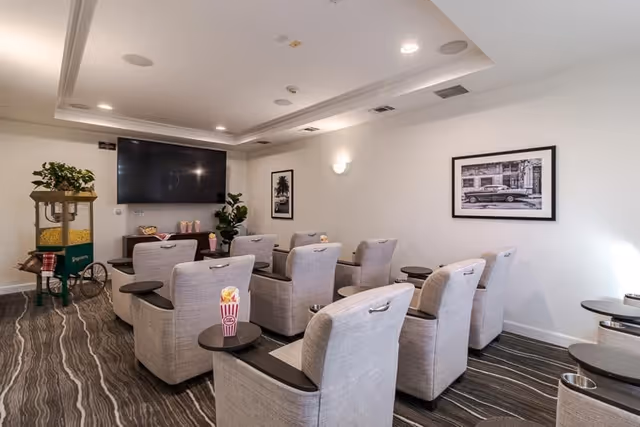 A small theater room with two rows of comfortable beige recliner chairs, each with a small attached table holding popcorn containers. A large flat-screen TV is mounted on the wall in front of the chairs. There is a popcorn machine on the left side of the room and framed black and white photographs on the walls. The room has a modern ceiling with recessed lighting and a patterned carpeted floor.