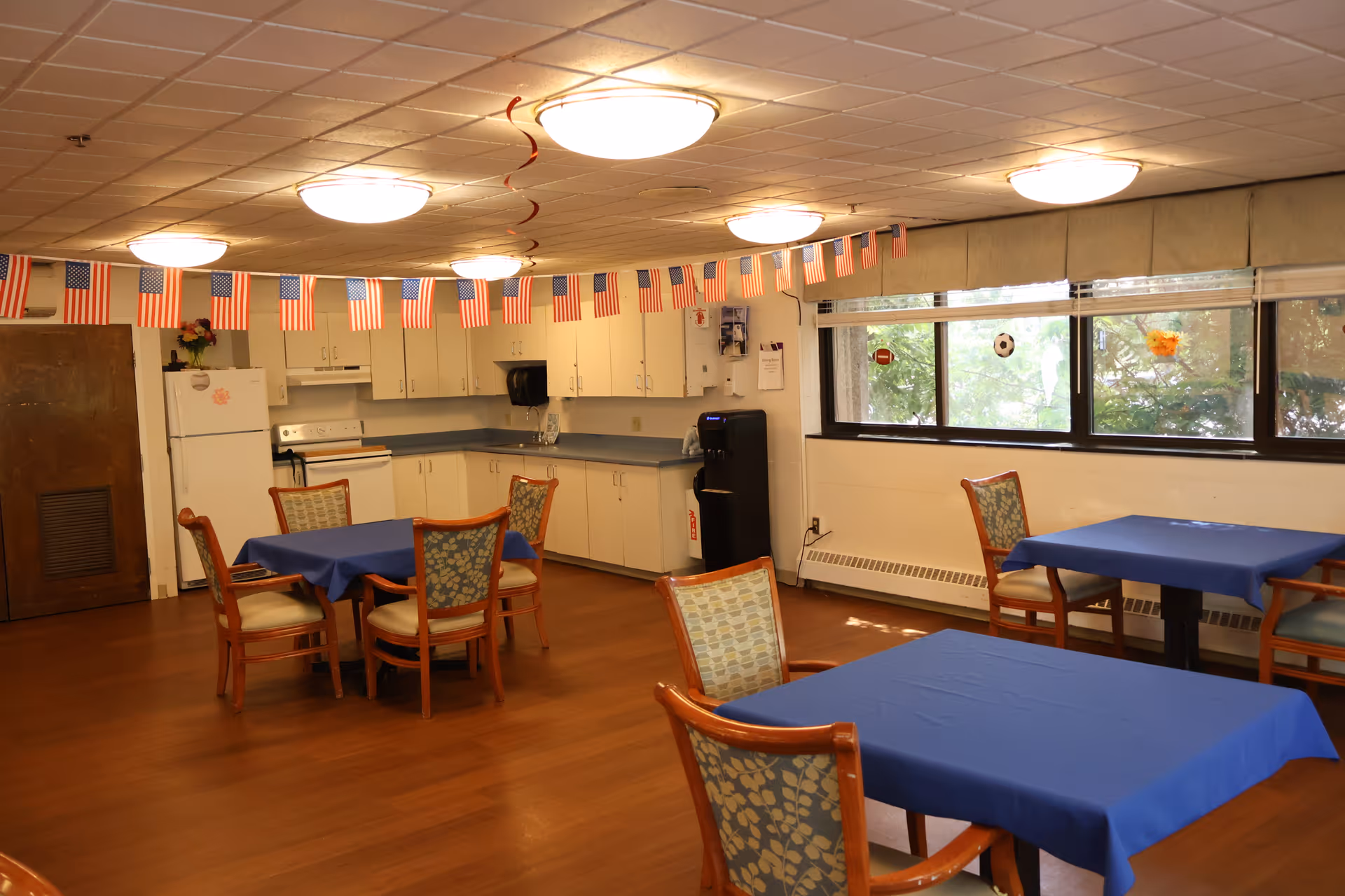 Communal dining room with blue-covered tables and chairs, a kitchenette along the back wall, and American flag bunting overhead.