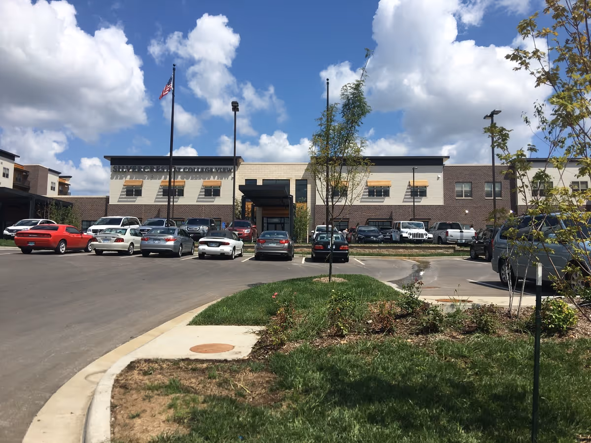 Front exterior view of Silvercrest at College View Senior Living facility with a parking lot in front, several parked cars, two flagpoles with an American flag, and a partly cloudy blue sky.
