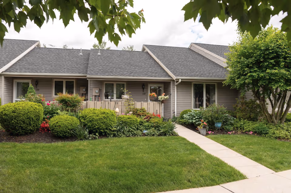 Exterior view of a single-story residential building with gray siding and a dark shingled roof. The building has windows and a small fenced patio area with hanging flower pots. In front of the building, there is a well-maintained lawn, various green shrubs, flowering plants, and a concrete walkway leading to the entrance. A tree with green leaves is visible on the right side, and tree branches partially frame the top of the image.