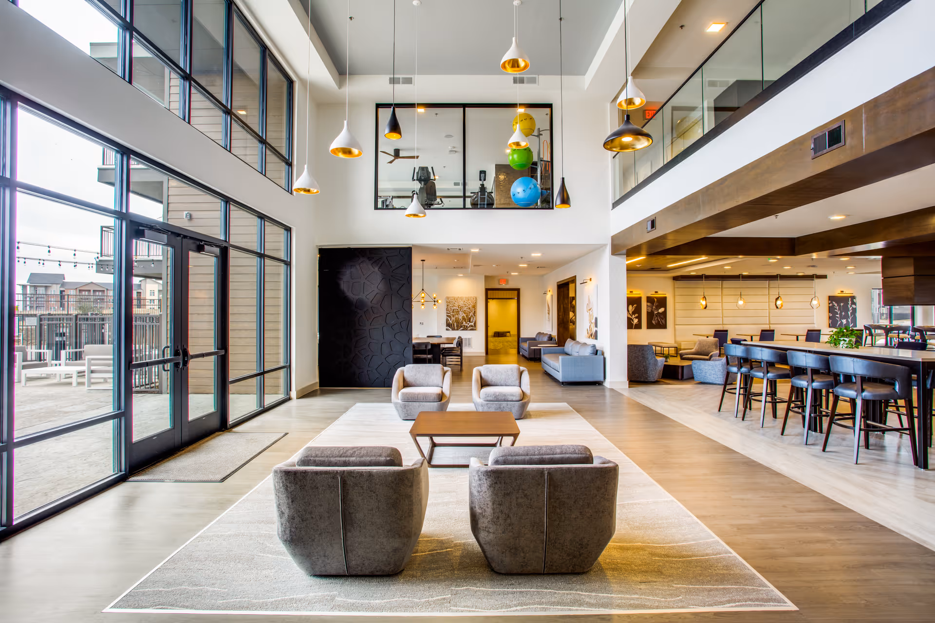 Spacious and modern common area with high ceilings and large windows allowing natural light. The room features a seating area with four gray armchairs around a wooden coffee table on a light rug. To the right, there is a long dining table with bar stools and additional seating areas with sofas and chairs. The upper level has a glass railing and exercise equipment visible through a window. Outside, there is a patio area with outdoor furniture.