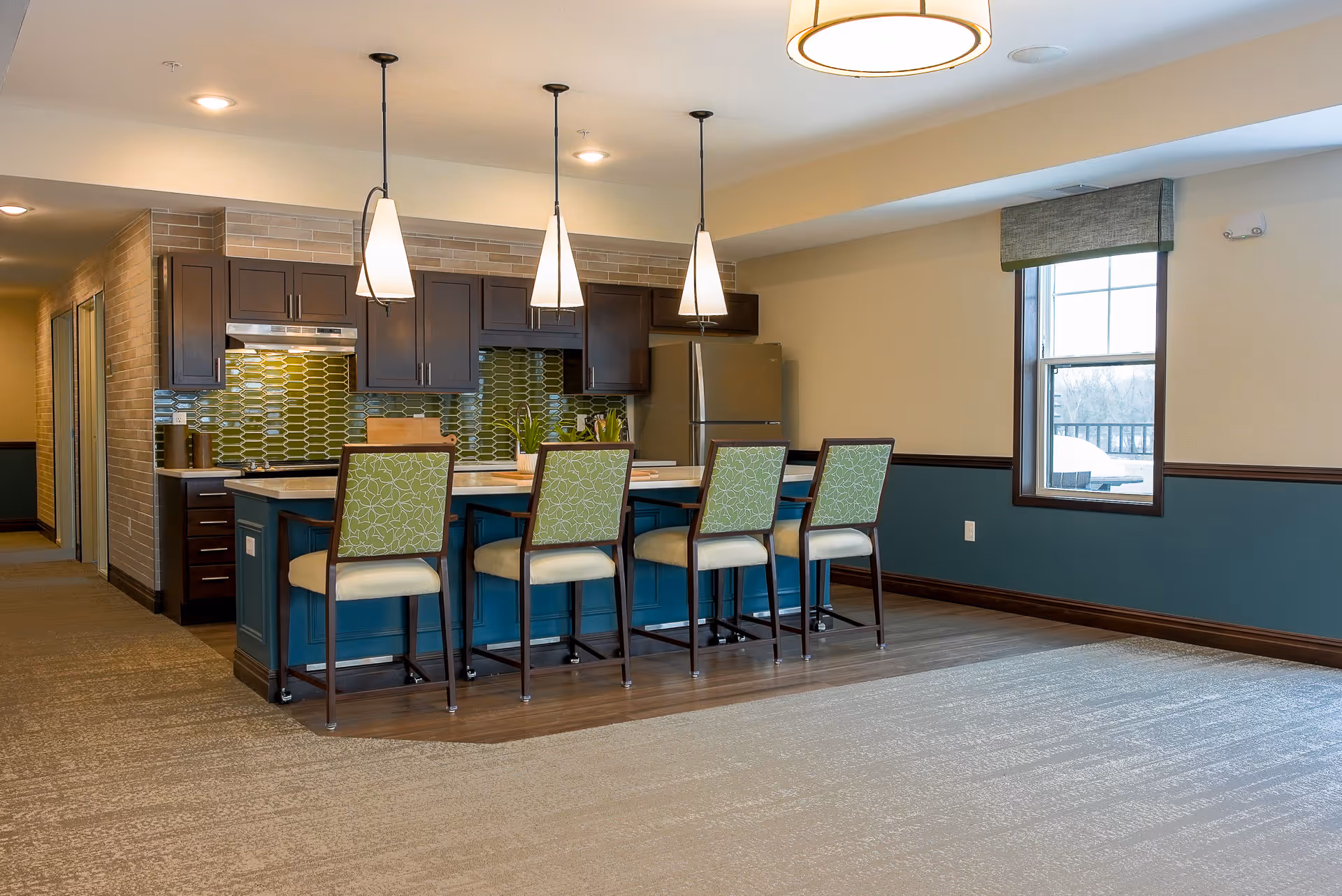 A modern kitchen area in a senior living facility with a blue island counter, four high chairs with green patterned backs, dark wood cabinets, a stainless steel refrigerator, and three pendant lights hanging above the island. There is a window on the right side letting in natural light.