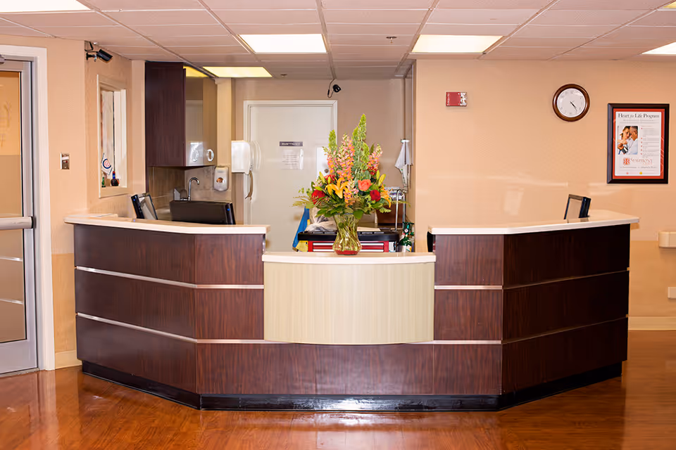 Reception desk area with a dark wood and light beige counter, a vase with colorful flowers on the counter, a clock and a framed poster on the beige wall behind, and a door and sink area in the background.