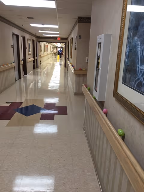 Long, glossy hallway inside a senior living facility with handrails, framed art, and a person visible at the far end.