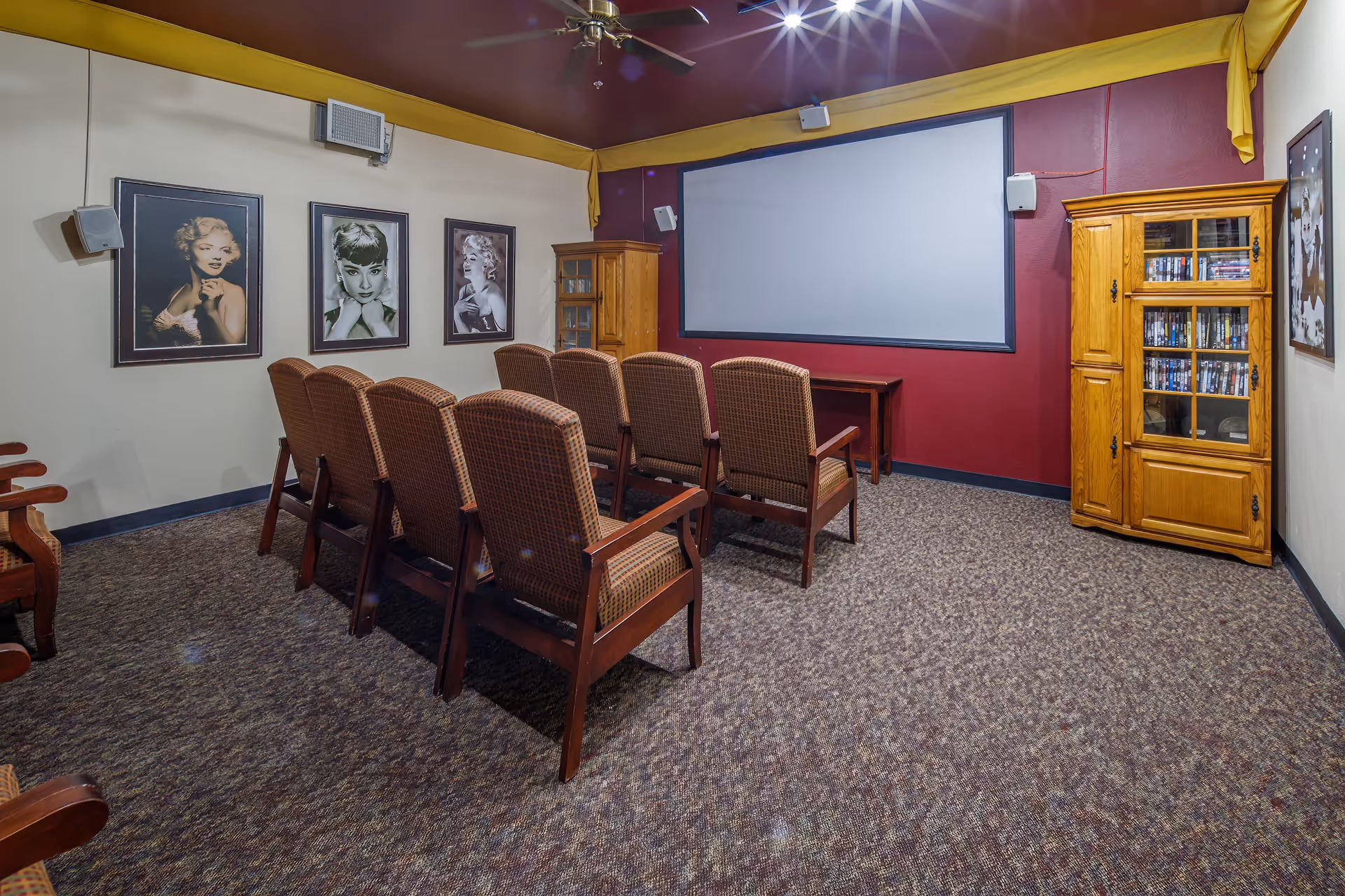 A small theater room with two rows of upholstered chairs facing a large blank projection screen. The walls are decorated with framed black and white portraits of classic Hollywood actresses. There are wooden cabinets on either side of the screen containing DVDs. The room has carpeted flooring and a ceiling fan with lights.
