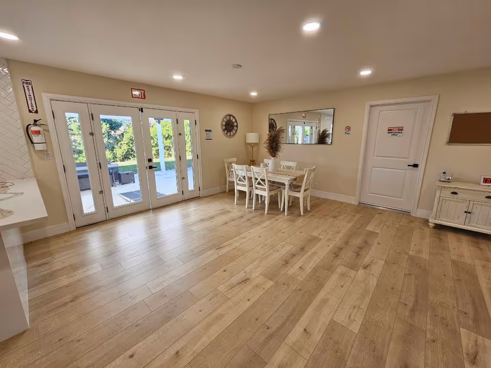 Bright communal room with wood flooring, a dining table and chairs near glass doors leading to an outdoor patio.