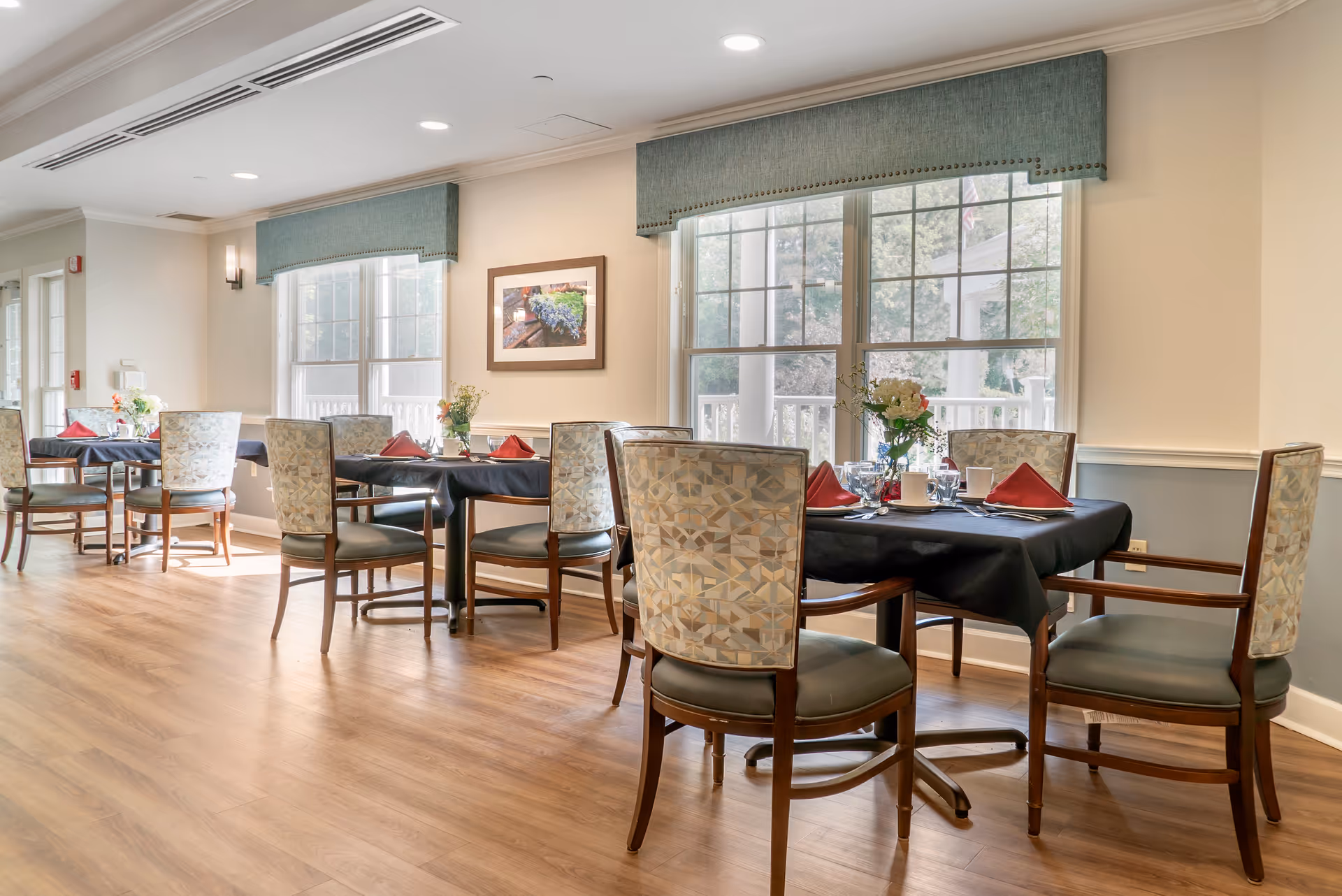 A bright dining room with wooden floors, several tables covered with black tablecloths, each set with red napkins, white cups, glasses, and floral centerpieces. Large windows with blue valances let in natural light, and a framed picture hangs on the wall.