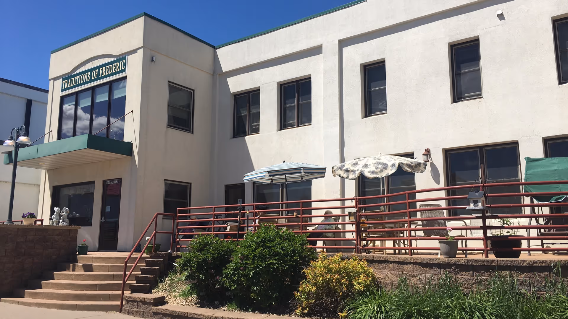 Exterior view of a two-story beige building with a green awning above the entrance. The building has a sign that reads 'Traditions of Frederic'. There is a patio area with red railings, outdoor chairs, tables, and umbrellas. Some greenery and bushes are visible in front of the patio under a clear blue sky.