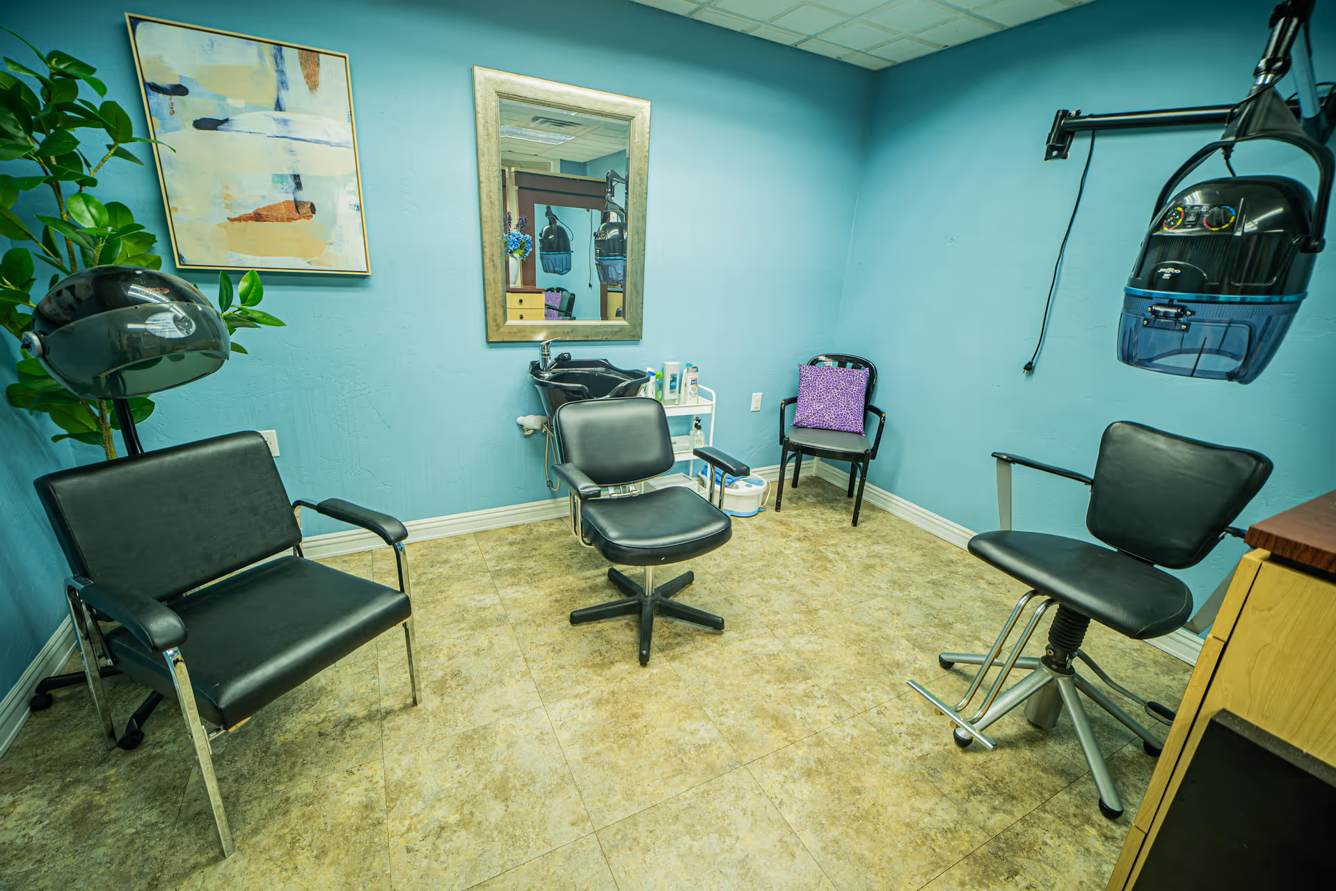 A small salon room with three black salon chairs, a hair washing station with a mirror above it, a hair dryer hood, a small side table with hair care products, a purple cushion on a chair in the corner, and a framed abstract painting on a teal wall.