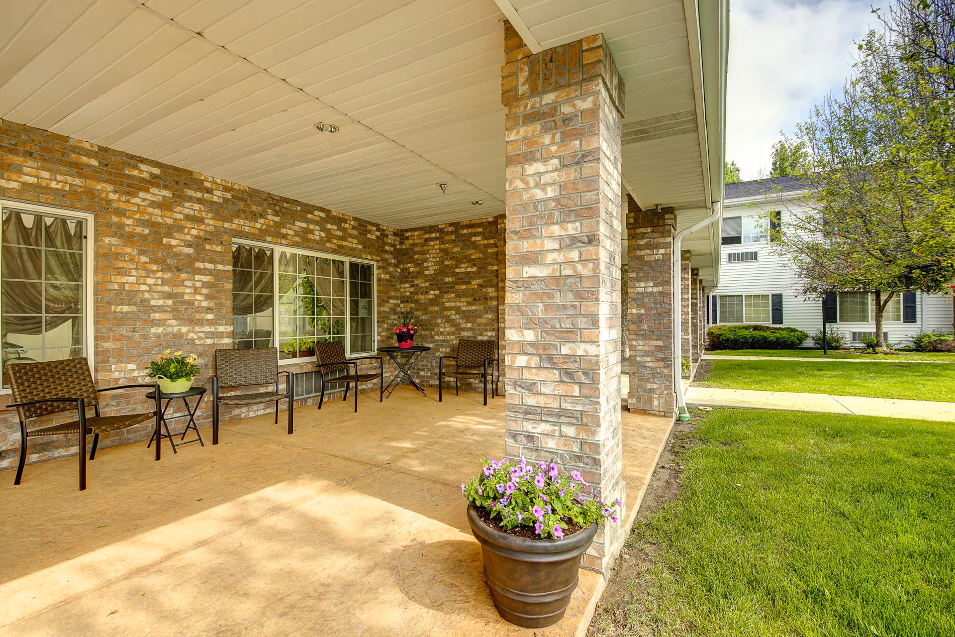 Covered outdoor patio area with brick pillars and walls, several brown wicker chairs and small tables with potted plants, adjacent to a grassy lawn and trees, with a white building visible in the background.
