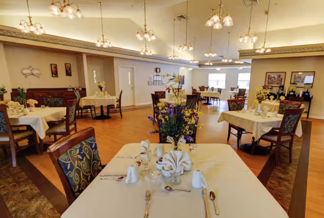 Dining room with neatly set tables, floral centerpieces, patterned chairs, and hanging chandeliers.
