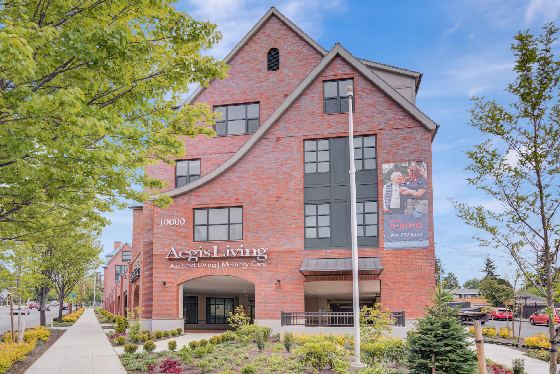 Exterior view of Aegis Living Greenwood, a red brick assisted living and memory care facility with multiple windows, a landscaped garden, and a sidewalk lined with trees. A banner on the building advertises job openings with an image of two women, one elderly and one younger.