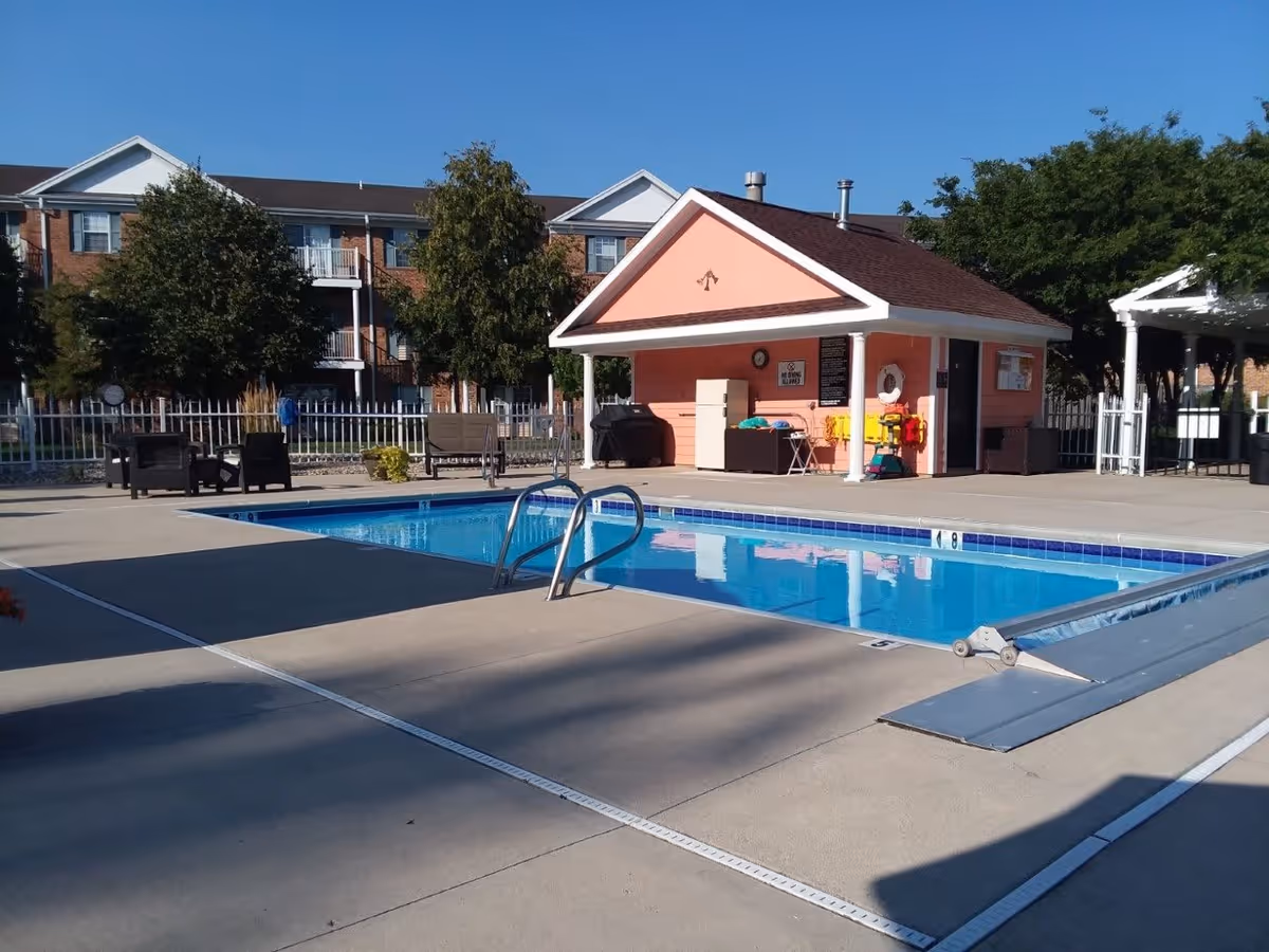 Outdoor swimming pool area at Georgetowne Place with a small pink pool house, poolside chairs, and a clear blue sky. There are trees and a multi-story brick building in the background.