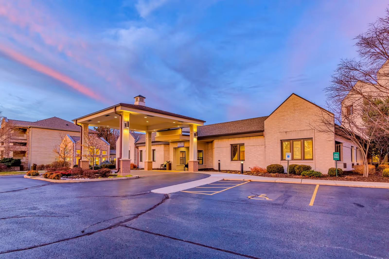 Exterior view of Harbour Village facility at dusk with a covered entrance, parking spaces including a handicapped spot, landscaped bushes, and a colorful sky with pink and blue clouds.