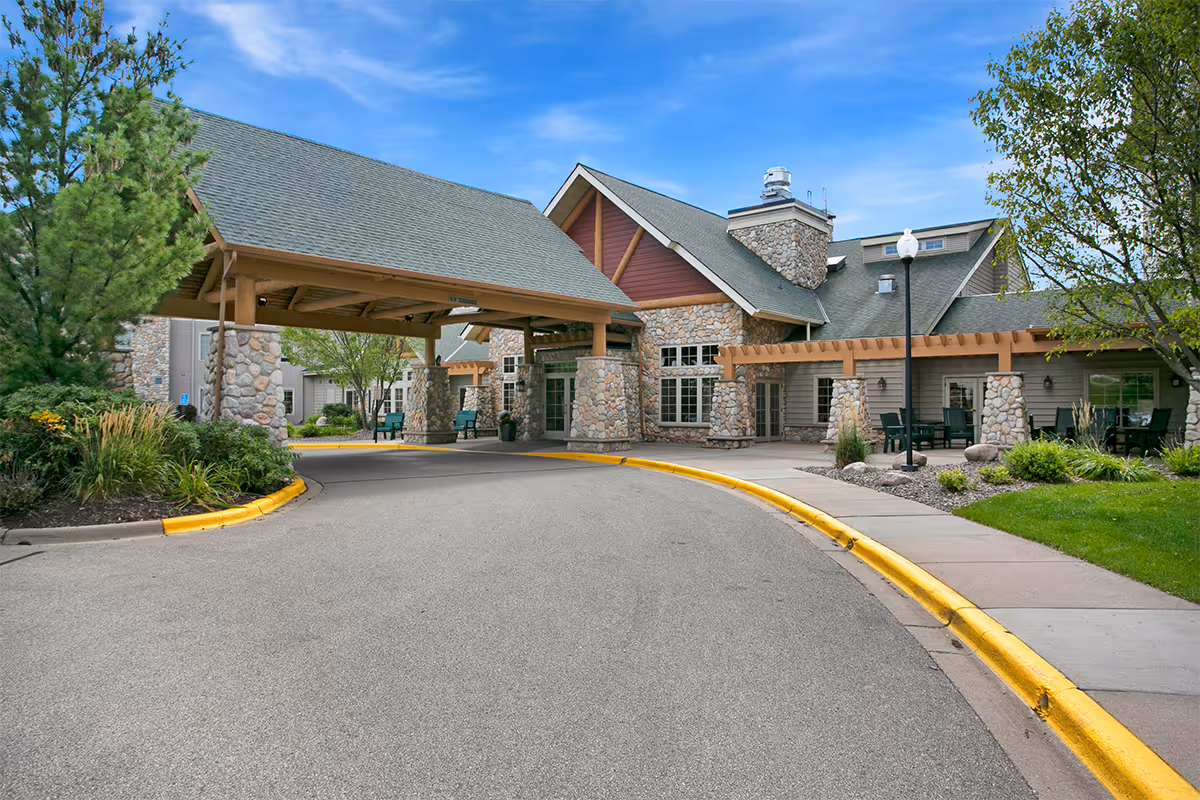 Front entrance of a senior living facility with a covered porte-cochère, stone facade, landscaped beds, and a curved driveway.