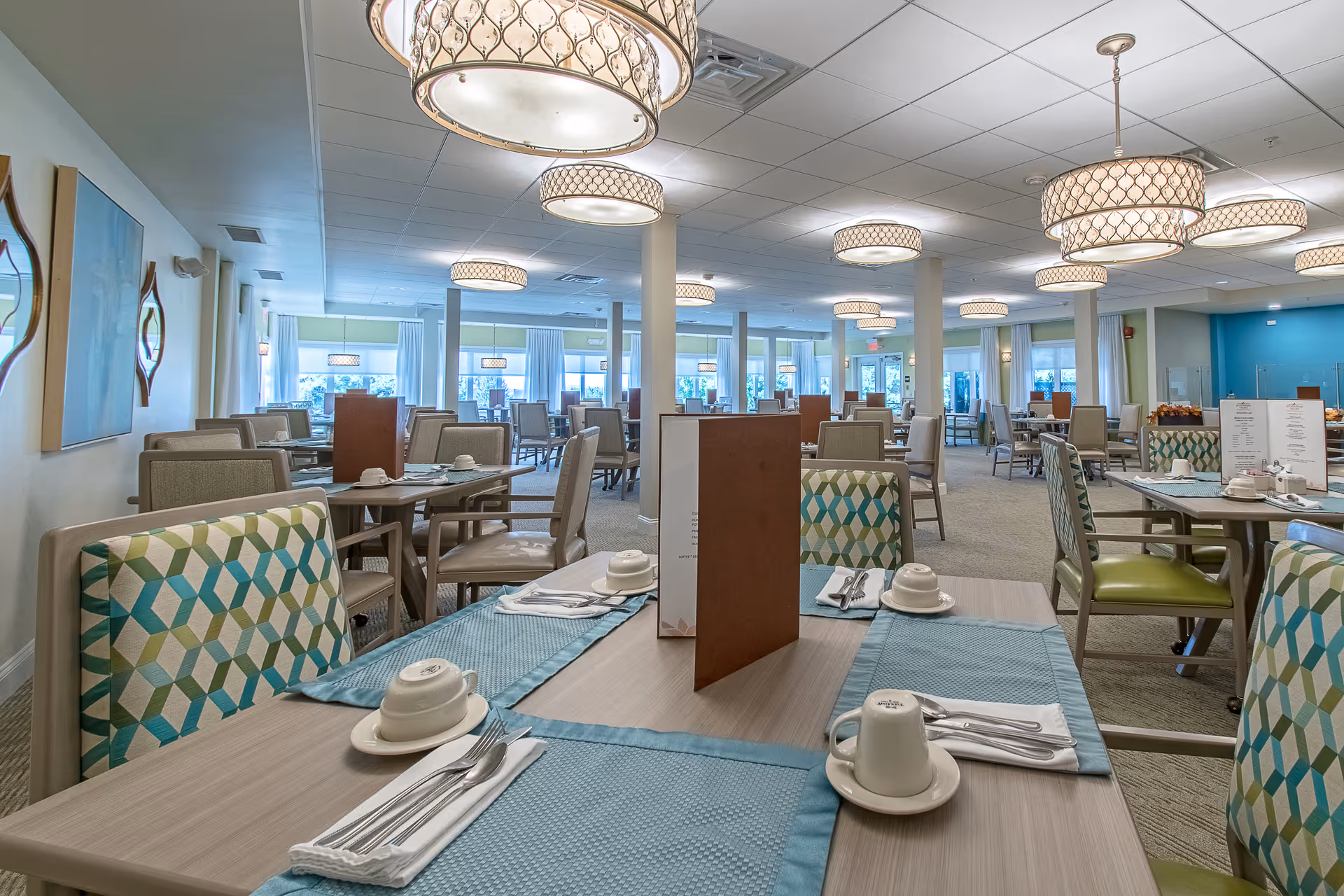 A spacious dining room with multiple tables set with blue placemats, white cups and saucers, silverware, and menus. The room features patterned chairs, large windows with white curtains, and modern circular chandeliers hanging from the ceiling.