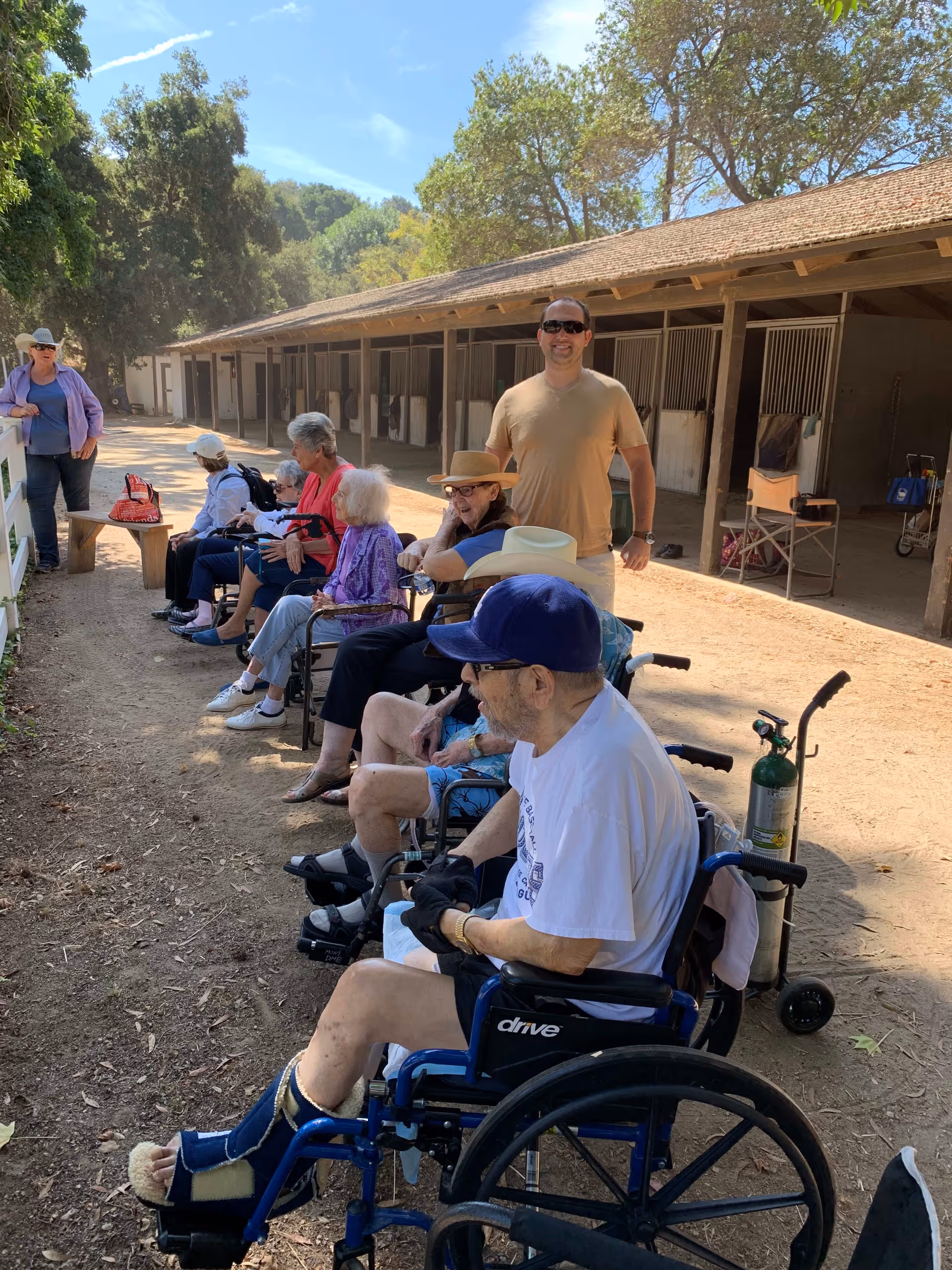 A group of elderly people, some in wheelchairs, sitting outdoors along a dirt path near a stable or barn area with horse stalls. A man in a tan shirt and sunglasses stands behind them, and another woman stands near a white fence. Trees and a clear blue sky are visible in the background.