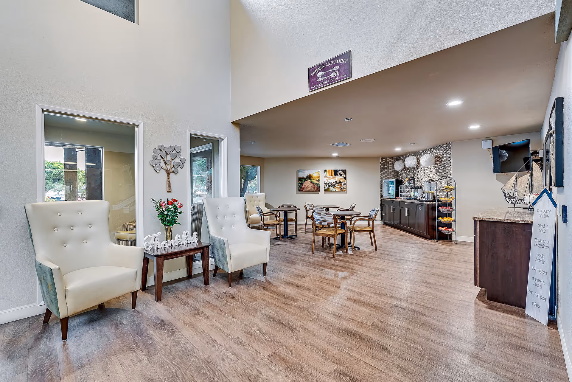 A senior living facility common area with two cream-colored armchairs and a small table with a flower vase and a 'Thankful' sign. In the background, there are several round tables with chairs, a refreshment station with drinks and snacks, and wall decorations including a sign that reads 'Friends and Family gather here'. The room has wood flooring and neutral-colored walls.