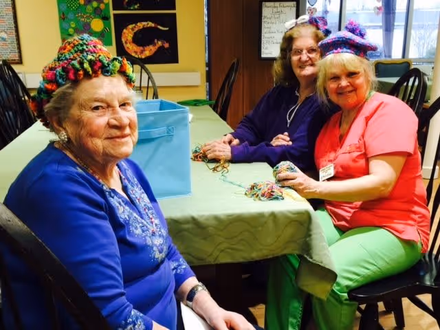 Three elderly women sitting around a table in a room, two of them wearing colorful knitted hats and smiling. They appear to be engaged in a knitting or craft activity with yarn on the table. The room has artwork on the walls and large windows letting in natural light.