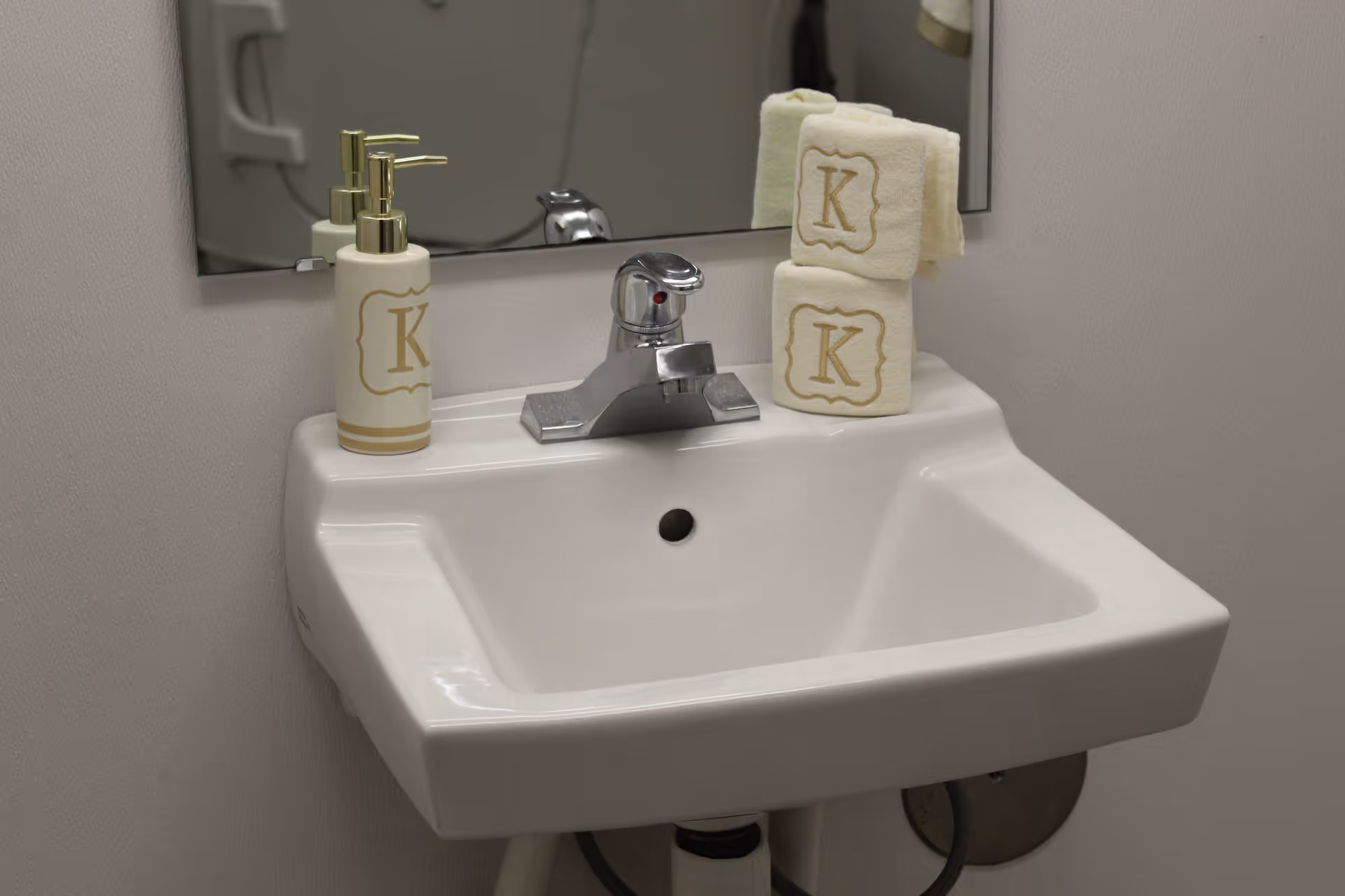 A white bathroom sink with a chrome faucet, a soap dispenser, and two folded towels with the letter 'K' embroidered on them, placed in front of a mirror.