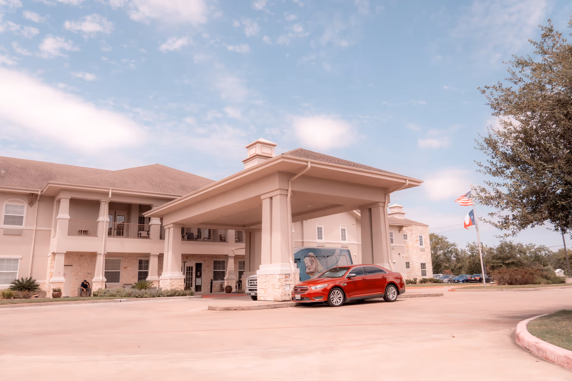 Exterior view of The Legacy at Falcon Point senior living facility showing the main entrance with a covered drop-off area. A red car is parked under the canopy, and an American flag and Texas state flag are visible on flagpoles to the right. The building has beige walls, stone accents, and a two-story design with balconies.