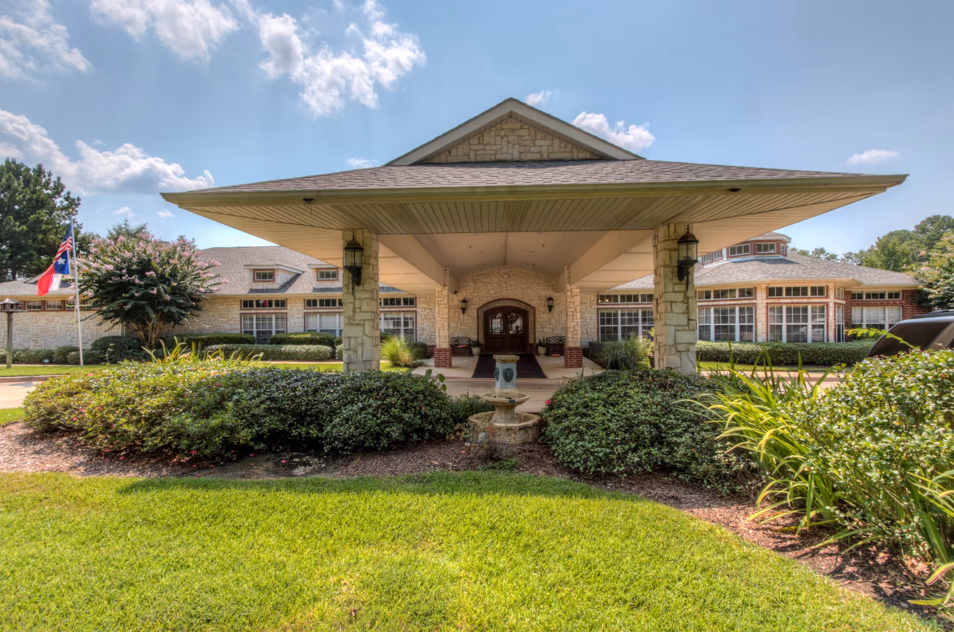 Front exterior view of Arabella of Kilgore Senior Living facility featuring a covered entrance with stone pillars, well-maintained landscaping with green bushes and grass, a small fountain in the center, and a clear blue sky with some clouds.