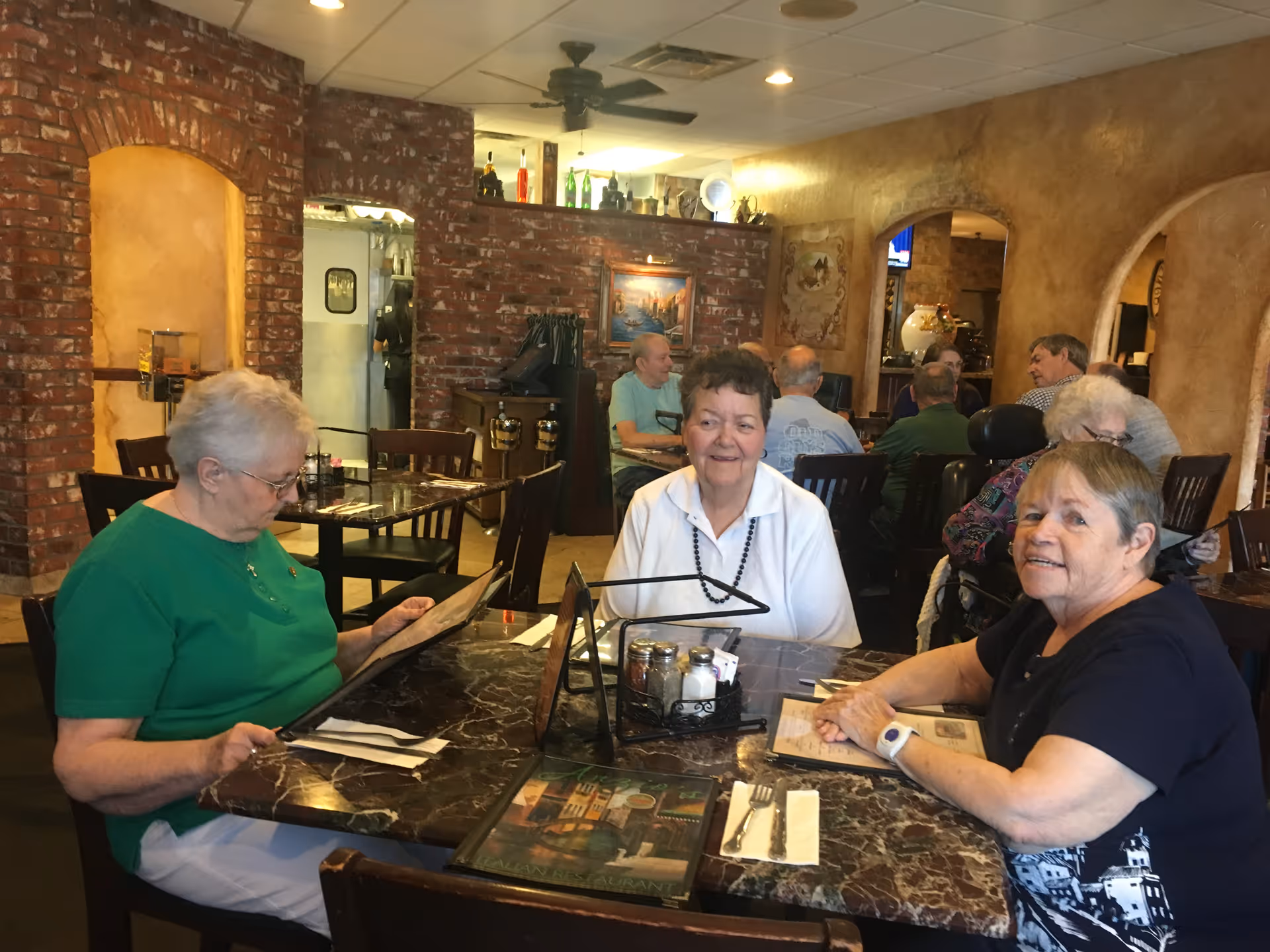 Three elderly women sitting at a marble table in a restaurant, two of them looking at menus while the third smiles at the camera. The restaurant has brick and stucco walls with arches and other diners visible in the background.